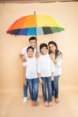 Happy indian family standing under big multicolor or colorful umbrella isolated on beige...