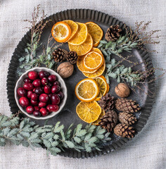 Weihnachtsdeko auf einem Teller aus Metall, eine Schale mit Cranberrys, getrocknete Orangenscheiben, Zapfen, Nüsse und Mimosenzweig, dekorieren im Advent