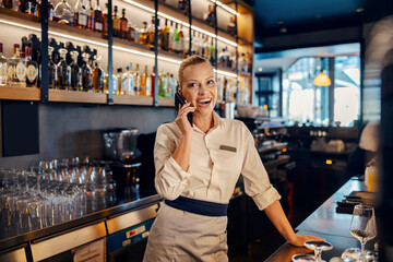 A happy female bartender is leaning on a counter and having a phone conversation in a bar.