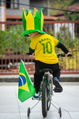 A happy kid smiling while riding a bike with the colors of the brazilian flag. He is using the Brazil National Team shirt celebrating the victory in some game on the World Cup of football (soccer).