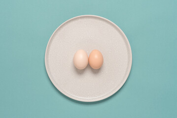 Two chicken light brown eggs on a beige plate, turquoise pastel background. Creative layout. The concept of minimalism and easter. Top view, flat lay.