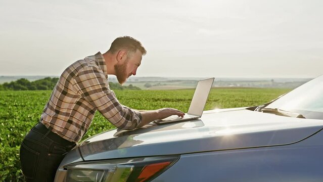A farmer working on a laptop on the hood of a car standing in the middle of a field. He turned and looked at the field.