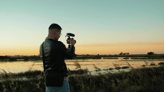 Attractive male videographer shooting a b-roll next to a beautiful lake during golden hour at sunset. Slow motion medium shot of an elegant young content creator wearing glasses and capturing video.