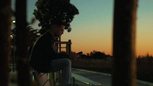 Professional drone pilot and videographer flying his drone and capturing social media video content for his clients. Slow motion of a young content creator wearing glasses sitting on a bench at dusk.