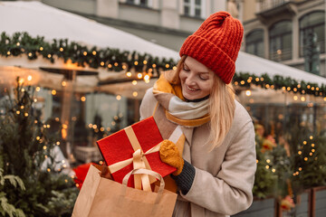 Fototapeta na wymiar Happy young winter woman with christmas gift. Gift Box Outdoors. Surprise. Cheerful attractive girl shopper with paper bags and presents. Christmas shopping in mall. Winter sales