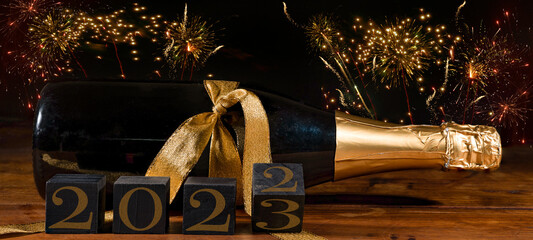 New Year 2022 / 2023 Sylvester New Year's Eve celebration holiday greeting card banner - Black cubes with year on sparkling wine or champagne bottle on table and firework in the background
