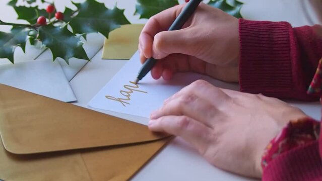Close up on the hands of a woman handwriting winter season's greetings on a christmas card, in gold ink cursive. A golden envelope is waiting on the desk, also decorated with a branch of holly 