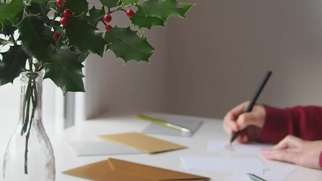 Closeup shot of a woman at her desk with holly decoration, preparing winter holidays by writing calligraphy cards to go with christmas presents