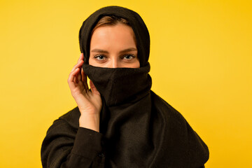 Adorable modern eastern woman wearing burqa looking at camera over isolated yellow background 