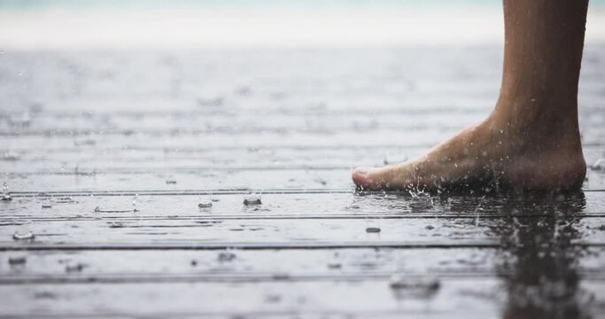 Women's barefoot covered in droplets of heavy rain in close-up. Caucasian Female legs walking on the wet wooden floor in rain weather. Happiness calmness concept. Slow motion