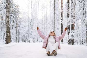 Young woman with blonde hair in winter clothes having fun sitting in snowy winter forest