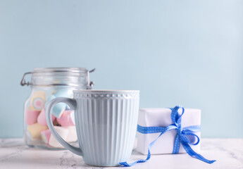 Festive postcard. Blue cup with hot drink, box with present and jar with colorful marshmallows on white marble background against blue wall. Selective focus. Place for text.