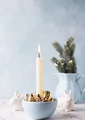 Foto auf Leinwand White burning candle, golden decorative cones, white bunnies and branches of Christmas tree in b pitcher on white marble background against blue  textured wall. Rabbit is symbool of 2023..  © daffodilred