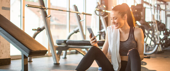 Cheerful sporty woman using phone after fitness workout in gym. Young smiling woman using phone during the break in gym. - obrazy, fototapety, plakaty