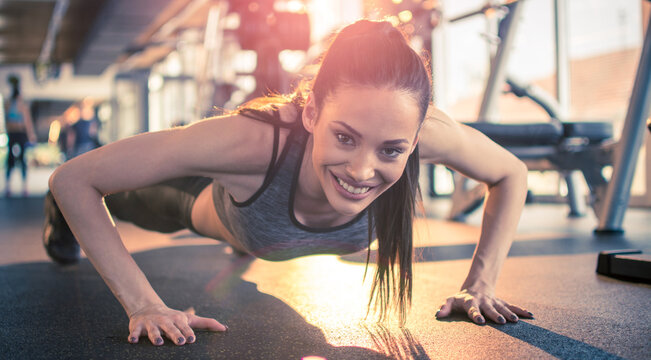 Fitness young woman doing push ups and looking at camera at gym. Friendly attractive girl doing push ups in front of sunlight at gym in the morning.