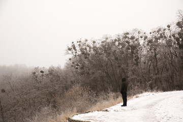 Obraz na płótnie Canvas A pensive man in black clothes stands on a high cliff near a river in winter