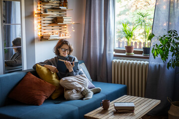 Young smiling cheerful woman in a warm sweater and eyeglasses reading a book while sitting on the couch in the room - obrazy, fototapety, plakaty