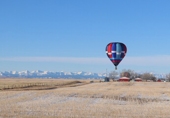 Blue, red and white hot air balloon floats peacefully above a red barn with the snow capped Rocky Mountains in the distant background of this winter landscape.