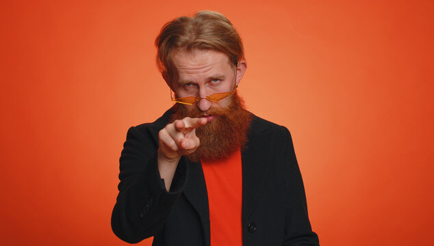 I am watching you. Blonde man in black jacket pointing at his eyes and camera, show I am watching you gesture, spying on someone. Young redhead guy boy isolated alone on orange studio wall background