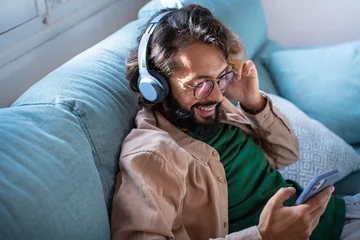 Poster Smiling young man wearing headphones listen to mobile music playing in smartphone app, happy guy relaxing holding using phone enjoy favorite songs relaxing sit on sofa at home. High quality photo © deliris