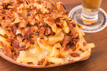 portion of french fries with cheese and bacon served on a table with a glass of beer blurred in the background