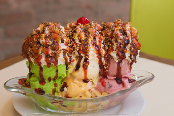 pistachio, cream and strawberry ice cream served in a glass dish topped with strawberry, chocolate, flakes, cherry, on the table with a blurred background of a brick wall