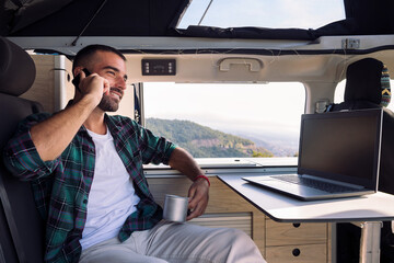 smiling young man working with a laptop inside his camper van and talking on mobile phone, concept of freedom and digital nomad lifestyle