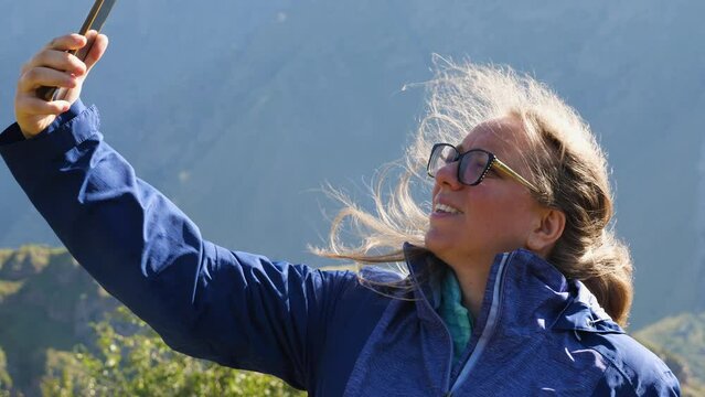 A girl with glasses is taking pictures of herself on her phone, her blond hair rippling in the wind. A woman tourist stands in front of mountains and takes selfies. Her hair is illuminated by sun