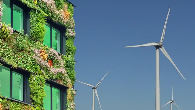 Pan of green sustainable building covered with blooming vertical hanging plants in front of rotating wind turbines