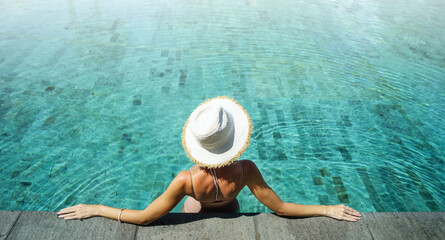 Back view of young woman wearing summer hat relaxing in big swimming pool with blue water on a sunny day.