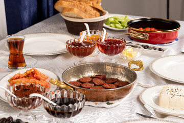 Breakfast table, morning family routine rich breakfast table. Turkish sausage called sucuk in frying pan ready to eat. Bread, cheese, tomatoes, jam, honey, tea in glass. Homemade dining table.