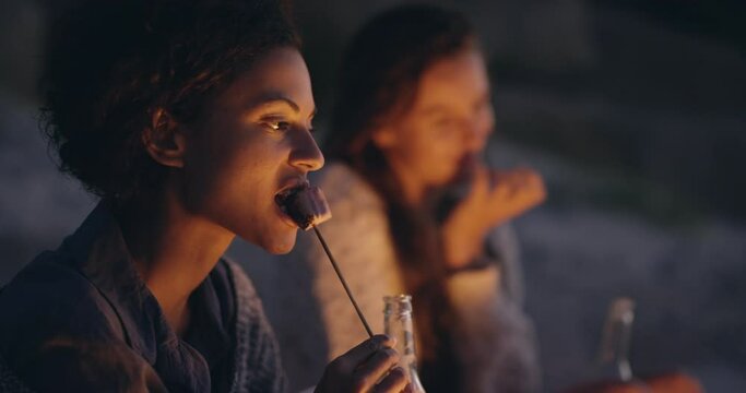 Woman, friends and night with fire, marshmallow and eating together for camping, bonding and relax. Women, diversity and toast candy at beach, dark and vacation with beer, drinking or social campfire