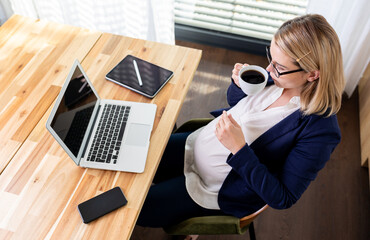 Pregnant woman at work drinking coffee with laptop tablet and smartphone lying on her desk