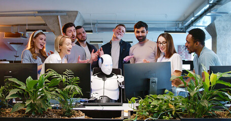 Robot sitting at table and typing on keyboard. People applauding. Cyborg working at computer in coworking space. Artificial intelligence, developers, Humanoid IT worker. Multiethnic team cheering up.