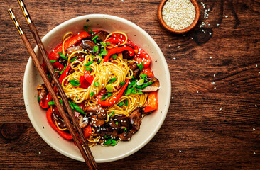 Vegan Stir fry noodles with vegetables, paprika, mushrooms, chives and sesame seeds in bowl. Wooden table background, top view, copy space