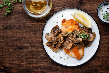 Fried chicken breast fillet with champignon mushrooms and onion in white wine sauce with thyme on plate, rustic wooden table background, top view