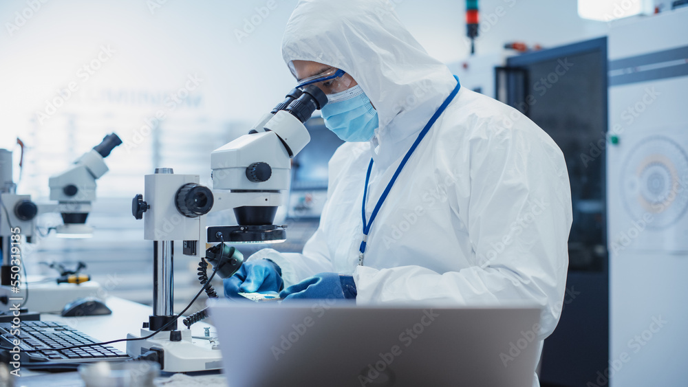 Poster Electronic Manufacturing Factory: Engineer in Sterile Coverall Examining a Circuit Board with Microchips Through a Microscope. Specialist Comparing Results on Laptop Computer