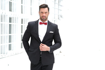 Handsome brunet in black suit with red bow tie on industrial building background.