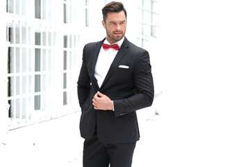 Handsome brunet in black suit with red bow tie on industrial building background.