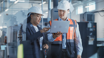 Two Professional Heavy Industry Engineers Wearing Safety Uniform and Hard Hats Discussing Factory Work on Laptop Computer. African American Specialist and Asian Technician at Work.