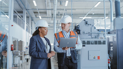 Two Professional Heavy Industry Engineers Walking at a Factory in Safety Uniform and Hard Hats, Discussing Work on Laptop Computer. Asian Specialist and African American Technician at Work.