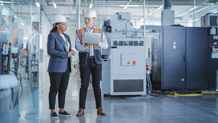 Two Professional Heavy Industry Colleagues in Hard Hats at Factory. Checking and Discussing Industrial Facility, Working on Laptop Computer. African American Engineer and Asian Technician at Work.