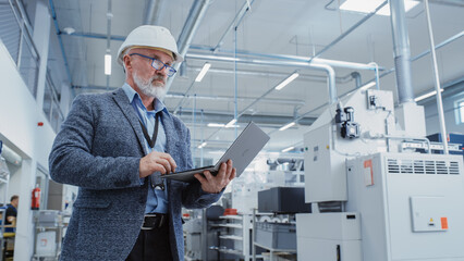 Portrait of a Bearded Middle Aged Chief Engineer Standing in a Factory Facility, Wearing Casual Suit and a White Hard Hat. Heavy Industry Specialist Working on Laptop Computer.