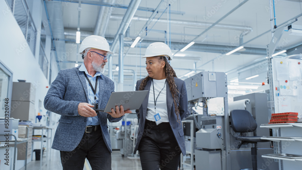 Poster portrait of a two diverse heavy industry engineers in hard hats walking with laptop computer and tal