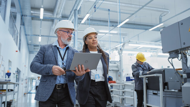 Two Professional Heavy Industry Engineers Wearing Hard Hats At Factory. Walking And Discussing Industrial Machine Facility, Working On Laptop. African American Manager And Technician At Work.