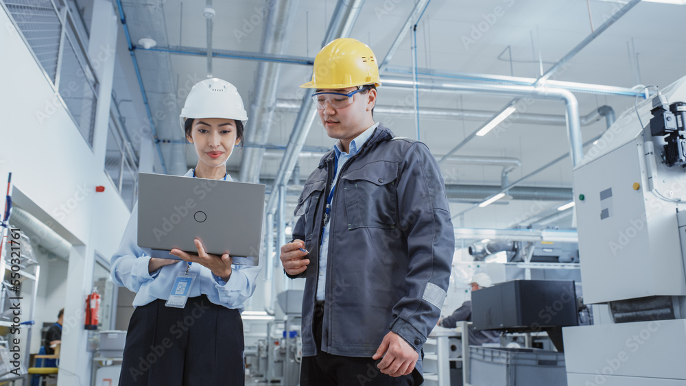 Poster two heavy industry employees in safety uniform and hard hats walking and talking in a factory. asian