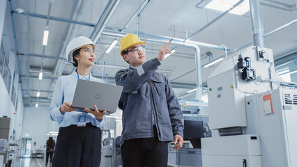 Two Heavy Industry Employees in Hard Hats Discussing Job Assignments at the Factory, Using Laptop Computer. Asian Engineer and Technician at Work Checking Safety Procedures.