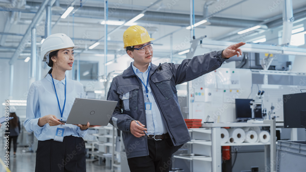 Poster two heavy industry employees in safety uniform and hard hats walking and talking in a factory. asian