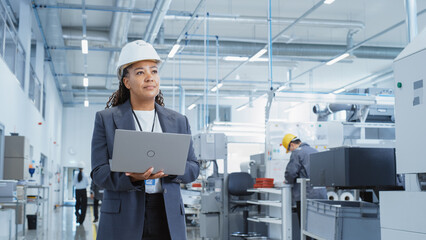 Portrait of a Happy Black Female Engineer in Hard Hat Standing and Using Laptop Computer at Electronic Manufacturing Factory. Technician Thinking About Daily Tasks and Working on Development Pipeline.