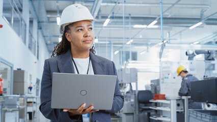 Black African Female Engineer in Hard Hat Standing and Using Laptop Computer at Electronic Manufacturing Factory. Technician Working on Daily Tasks and Research and Development Data.